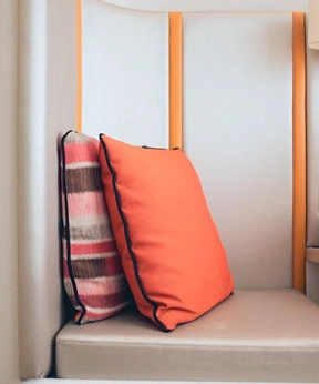Modern döşemeli bir sandalyenin üzerine yerleştirilen parlak turuncu bir minder, minimalist bir iç mekana sıcak bir dokunuş katıyor.